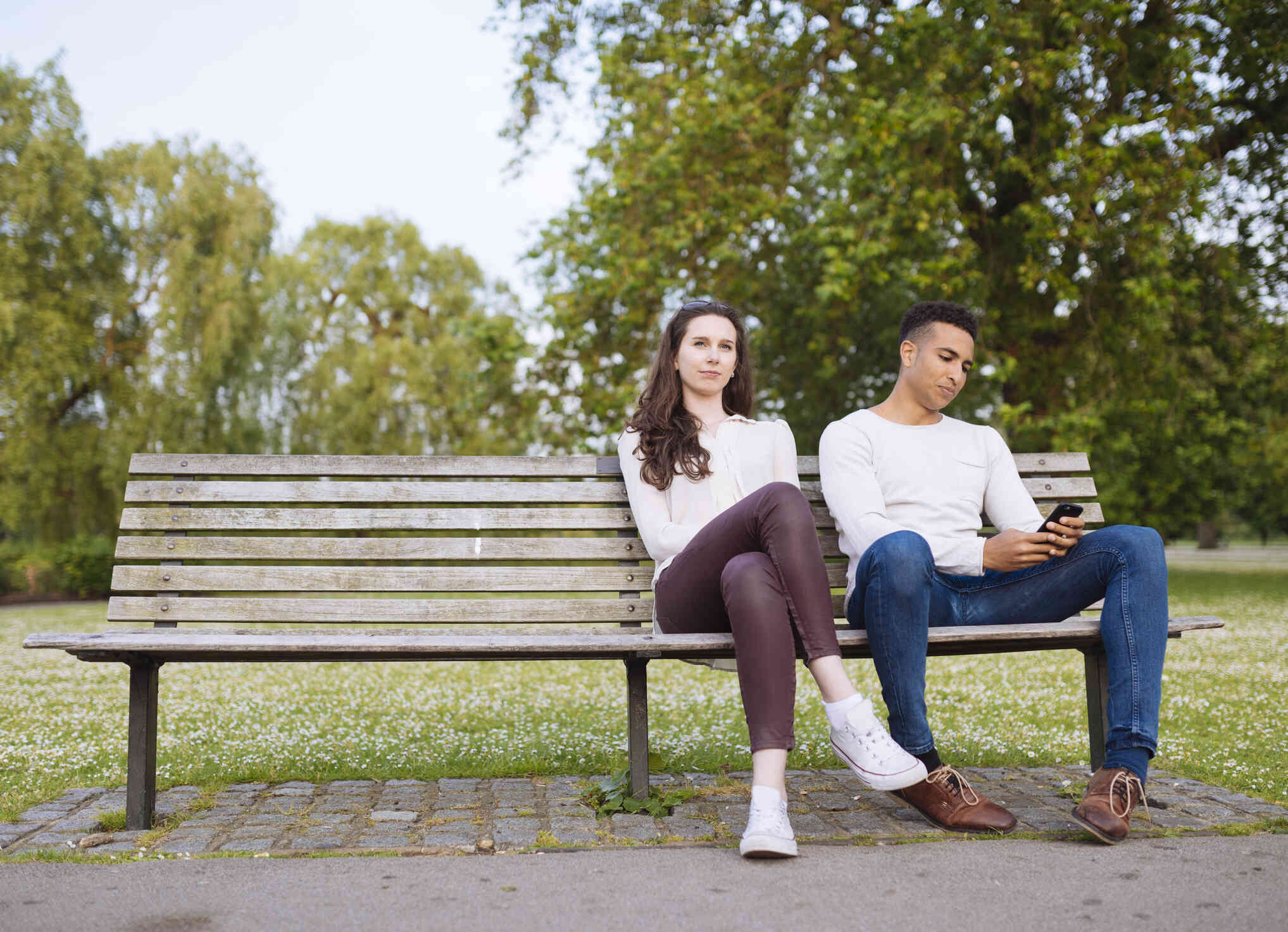 A male and female couple sit next to each other on a park bench on a sunny day while looking upset.